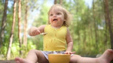 Kirli giysileri ve yüzü bebek maması meyvesi ya da sebze püresi yiyen küçük sevimli sarışın kız çocuğu yazın dışarıda sarı tabaktan kaşıkla yemek yiyor. Sağlıklı mutlu çocukluk kavramı.