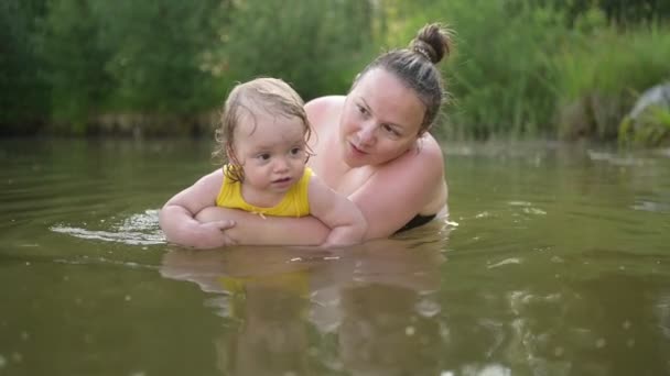Kleine lustige süße blonde Mädchen Kleinkind im gelben Body lacht lernt draußen am Sommersee schwimmen. Plus Größe Körper positiv lächelnde Frau Mutter Baby Schwimmen in natürlichen Pool. Wasserspritzer. — Stockvideo
