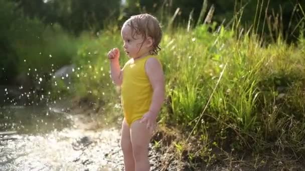 Lilla roliga söta blonda flicka barn småbarn i gul våt body suit spelar vid sjön stranden vid solnedgången utanför. Baby med lera på händer och ben. Vattenaktiviteter på sommaren. — Stockvideo