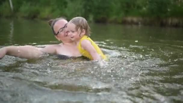 黄色のボディスーツの小さな面白いかわいいブロンドの女の子の子供の幼児笑いは夏の湖で外で泳ぐことを学びます。プラスサイズボディ肯定的な笑顔の女性の母親の赤ちゃんは自然のプールで泳ぐ。水が噴き出す. — ストック動画