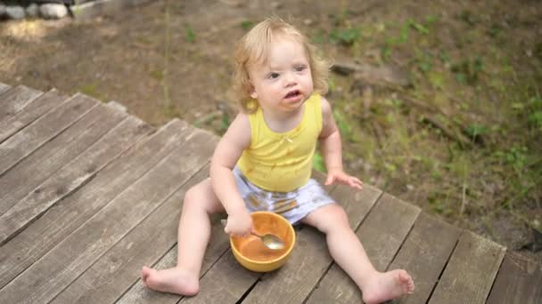 Klein grappig blond meisje kind peuter met vuile kleren en gezicht eten babyvoeding fruit of groente puree met lepel van gele plaat buiten in de zomer. Gezond concept voor een gelukkige jeugd. — Stockvideo