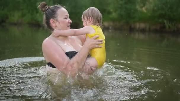 穿着黄色紧身衣笑着的可爱的金发小女孩在夏天的湖中学习游泳。加上身材瘦小、面带微笑的女人妈妈宝宝在自然游泳池里游泳。水花四溅. — 图库视频影像