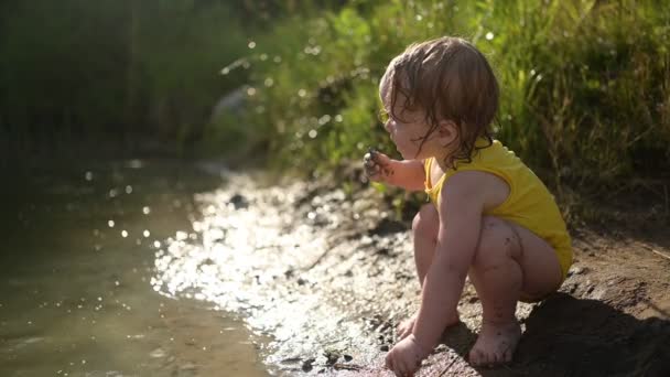 Μικρό αστείο χαριτωμένο ξανθό κορίτσι παιδί νήπιο σε κίτρινο υγρό κοστούμι του σώματος που παίζει δίπλα στην όχθη της λίμνης στο ηλιοβασίλεμα έξω. Μωρό με λάσπη στα χέρια και τα πόδια. Δραστηριότητες ύδρευσης το καλοκαίρι. — Αρχείο Βίντεο