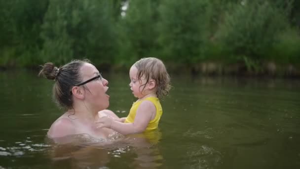 Pouco engraçado bonito menina loira criança criança em amarelo bodysuit rindo aprende a nadar fora no lago de verão. Além de corpo tamanho positivo sorridente mulher mãe bebê nadando na piscina natural. Salpicos de água. — Vídeo de Stock