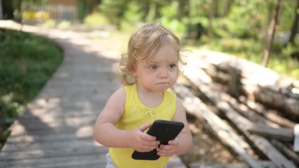 Little funny cute blonde girl child toddler holding playing big black smartphone outside at summer at countryside. Healthy happy childhood concept. Kids and technologies. — Stock Video