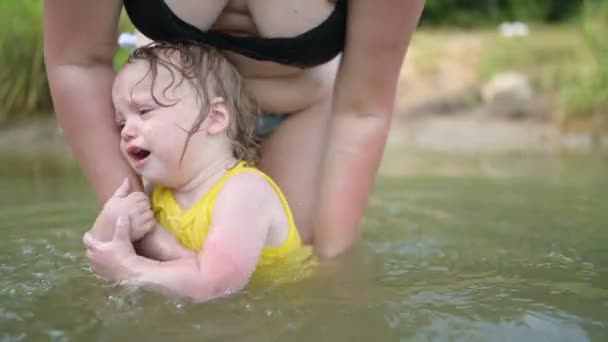Μικρό αστείο χαριτωμένο ξανθό κορίτσι παιδί νήπιο σε κίτρινο κοστούμι του σώματος κλαίει φοβούνται να μάθουν κολύμπι έξω στο καλοκαίρι λίμνη. Συν μέγεθος σώματος θετική γυναίκα μητέρα κολύμπι στη φυσική πισίνα. Παιδικό ξέσπασμα — Αρχείο Βίντεο