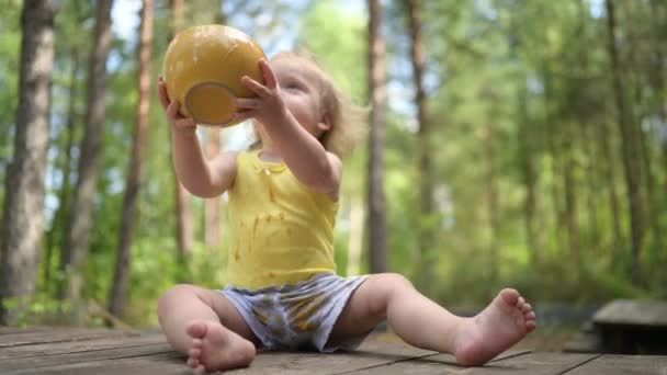 Kis vicces aranyos szőke lány gyermek kisgyermek piszkos ruhák és arc eszik baba élelmiszer gyümölcs vagy zöldség püré kanállal sárga tányéron kívül nyáron. Egészséges boldog gyermekkori koncepció. — Stock videók