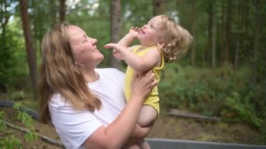 Annesinin kollarında bukleleri olan küçük tatlı bebek sarışın kız. Anne ve kızı yazın kırsal alanda ön bahçede birlikte vakit geçiriyorlar. Sağlıklı mutlu aile çocukluğu kavramı.