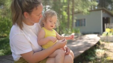 Annesinin kollarında bukleleri olan küçük tatlı bebek sarışın kız. Anne ve kızı yazın kırsal alanda ön bahçede birlikte vakit geçiriyorlar. Sağlıklı mutlu aile çocukluğu kavramı.