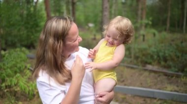 Annesinin kollarında bukleleri olan küçük tatlı bebek sarışın kız. Anne ve kızı yazın kırsal alanda ön bahçede birlikte vakit geçiriyorlar. Sağlıklı mutlu aile çocukluğu kavramı.