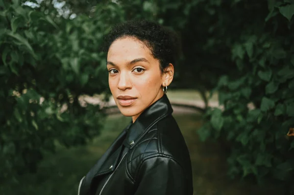 Fashion close up portrait of atraktive young natural beauty African American woman with afro hair in black skin jacket pospose in nature park in green foliage — Stok Foto