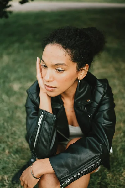 Potret busana wanita muda Amerika yang cantik dan percaya diri dengan rambut afro dalam jaket kulit hitam yang berpose di taman alam dalam dedaunan hijau — Stok Foto