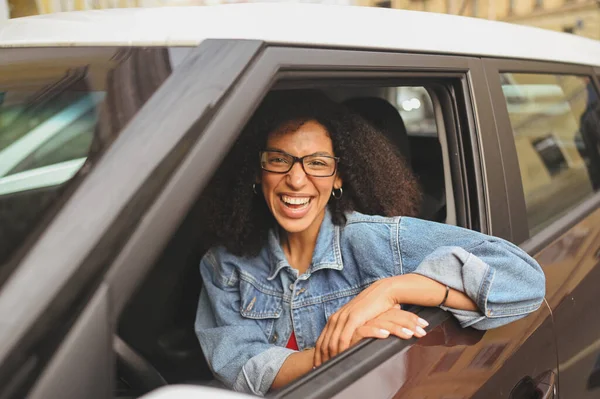 Happy young tersenyum muda Afrika Amerika wanita hitam berambut sopir dalam gelas duduk di mobil baru coklat, tersenyum melihat kamera menikmati perjalanan. Mengemudi kursus dan konsep asuransi jiwa — Stok Foto