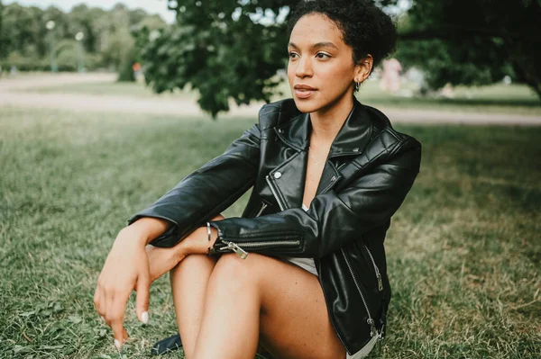 Retrato de moda de atractiva joven confiada belleza natural mujer afroamericana con pelo afro en chaqueta de cuero negro posando en el parque natural en follaje verde —  Fotos de Stock