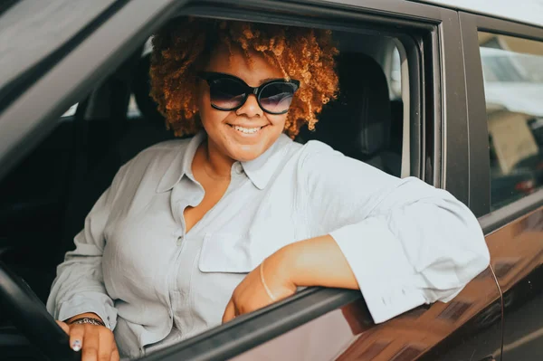 Jovem feliz sorrindo Africano americano mulher vermelho afro motorista de cabelos em óculos de sol sentado em novo carro marrom, sorrindo olhando para a câmera desfrutando viagem. Cursos de condução e seguro de vida — Fotografia de Stock
