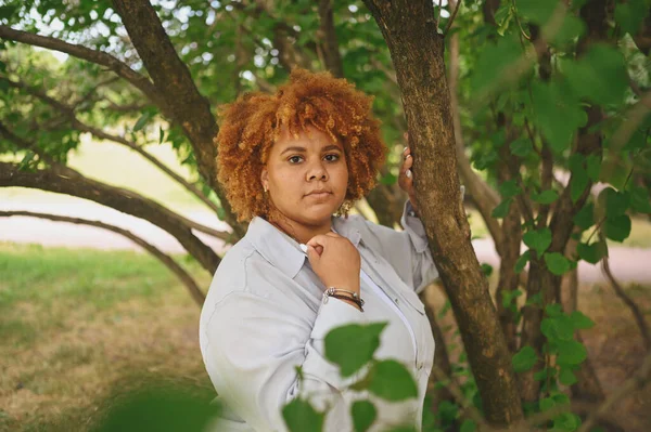 Fashion portrait of young naturally beautiful plus size African American woman in light trouser suit with afro red hair posing in nature parkland in green foliage. Diversity and body positive concept.