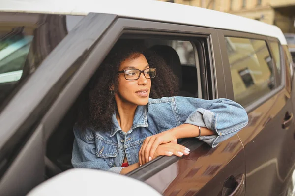 หญิงสาวชาวแอฟริกันอเมริกันที่มีความสุข คนขับรถผมดําในแว่นตานั่งอยู่ในรถสีน้ําตาลใหม่ยิ้มมองกล้องเพลิดเพลินกับการเดินทาง หลักสูตรการขับขี่และแนวคิดประกันชีวิต — ภาพถ่ายสต็อก