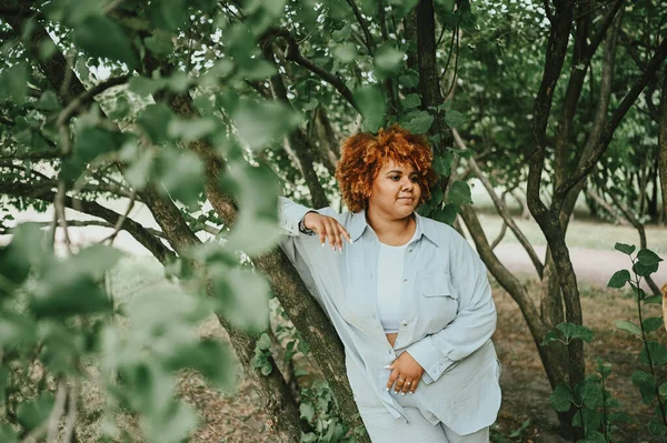 Potret mode wanita muda cantik secara alami ditambah ukuran wanita Afrika Amerika dengan pakaian celana panjang terang dengan rambut merah afro yang berpose di taman alam dalam dedaunan hijau. Perbedaan dan konsep positif tubuh. — Stok Foto