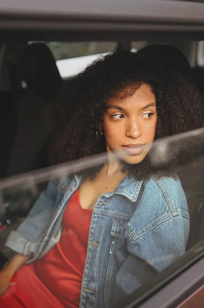 Cantik wanita muda Afrika Amerika berambut hitam driver dalam jaket jins duduk di mobil cokelat baru, menikmati perjalanan. Mengemudi kursus dan konsep asuransi jiwa — Stok Foto