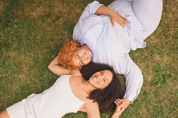 Pasangan muda Afrika-Amerika lesbian yang bahagia berbaring di rumput sambil tertawa sambil menjulurkan tangan sambil menutup kamera, pemandangan puncak musim panas. Konsep komunitas LGBT. Perempuan teman menikmati saat-saat cinta. — Stok Foto