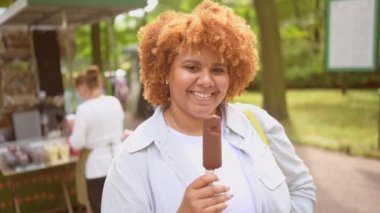 Doğal olarak güzel, gülümseyen ve Afro kızıl saçlı Afro-Amerikan bir kadının halk parkında dondurma yerken çekilmiş hayat tarzı portresi. Çeşitlilik ve beden pozitif konsept.