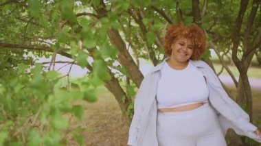 Doğuştan güzel, gülümseyen, Afro kızıl saçlı, yeşil yapraklı, Afro-Amerikan bir kadının yavaş çekim portresi. Çeşitlilik ve beden pozitif konsept.