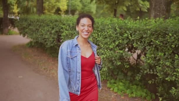 Retrato de movimento lento de moda sensual atraente jovem mulher afro-americana naturalmente bonita com cabelo preto na jaqueta de jeans e vestido vermelho andando no parque verde da natureza no verão. — Vídeo de Stock