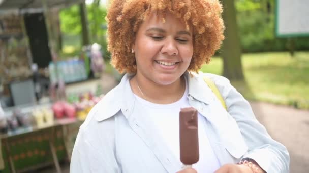 Lifestyle-Porträt einer lächelnden jungen, natürlich schönen Afroamerikanerin mit afroroten Haaren, die im öffentlichen Park Eis am Stiel aus Schokolade isst. Diversität und körperliches positives Konzept. — Stockvideo