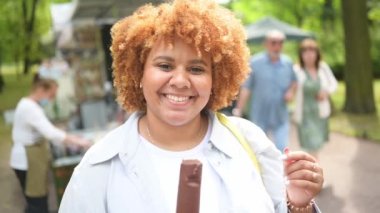 Doğal olarak güzel, gülümseyen ve Afro kızıl saçlı Afro-Amerikan bir kadının halk parkında dondurma yerken çekilmiş hayat tarzı portresi. Çeşitlilik ve beden pozitif konsept.