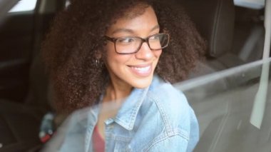Gülümseyen genç, Afro-Amerikan kadın siyah saçlı, gözlüklü, yeni kahverengi arabasında oturan, gülümseyen ve yolculuğun tadını çıkaran kameraya bakan. Sürüş dersleri ve hayat sigortası konsepti