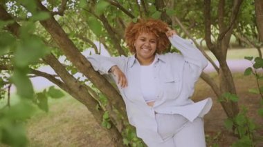 Doğuştan güzel, gülümseyen, Afro kızıl saçlı, yeşil yapraklı, Afro-Amerikan bir kadının yavaş çekim portresi. Çeşitlilik ve beden pozitif konsept.