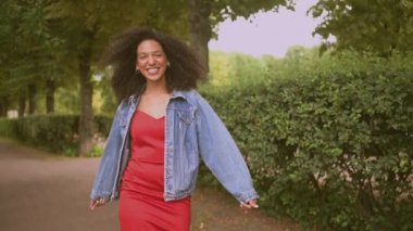 Şehvetli, çekici, doğuştan güzel, siyah saçlı, kot ceketli, kırmızı elbiseli, yaz aylarında yeşil doğa parkında yürüyen Afro-Amerikan kadının yavaş çekim portresi..