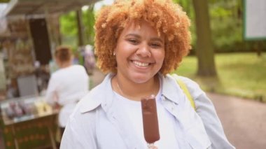 Doğal olarak güzel, gülümseyen ve Afro kızıl saçlı Afro-Amerikan bir kadının halk parkında dondurma yerken çekilmiş hayat tarzı portresi. Çeşitlilik ve beden pozitif konsept.