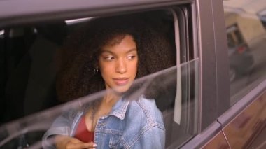 Gülümseyen genç, Afro-Amerikan kadın siyah saçlı, gözlüklü, yeni kahverengi arabasında oturan, gülümseyen ve yolculuğun tadını çıkaran kameraya bakan. Sürüş dersleri ve hayat sigortası konsepti