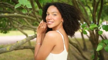 Moda, afro saçlı, yeşil yapraklarla kaplı doğa parkında poz veren çekici, doğuştan güzel Afro-Amerikan kadının yavaş çekim portresini kapatır..