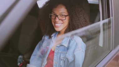 Gülümseyen genç, Afro-Amerikan kadın siyah saçlı, gözlüklü, yeni kahverengi arabasında oturan, gülümseyen ve yolculuğun tadını çıkaran kameraya bakan. Sürüş dersleri ve hayat sigortası konsepti