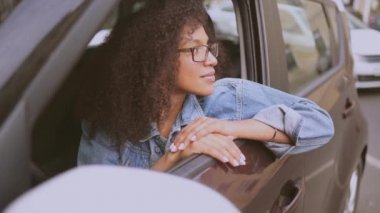Gülümseyen genç, Afro-Amerikan kadın siyah saçlı, gözlüklü, yeni kahverengi arabasında oturan, gülümseyen ve yolculuğun tadını çıkaran kameraya bakan. Sürüş dersleri ve hayat sigortası konsepti
