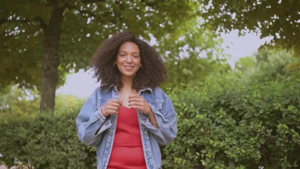 Moda cámara lenta retrato de sensual atractiva joven naturalmente hermosa mujer afroamericana con pelo negro en chaqueta vaqueros y vestido rojo caminando en verde naturaleza parque en verano. — Vídeos de Stock