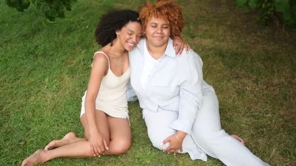 Young beautiful happy lesbian African American couple sitting on green grass hugging outside at nature summer park laughing closing from camera. LGBT community concept. Female friends love moments. — Stock Video