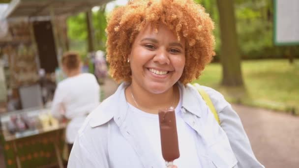 Styl życia portret uśmiechnięty młody naturalnie piękne plus size Afroamerykanka z afro rude włosy jedzenie lodów lodów czekoladowych lodów w parku publicznym. Różnorodność i pozytywna koncepcja ciała. — Wideo stockowe