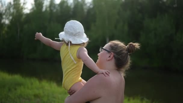 Kicsi aranyos kisbaba szőke lány, fürtökkel az anyai karján. Anya és lánya együtt játszanak a szabadban, az udvar előtt nyaranta. Egészséges boldog családi gyermekkori koncepció. — Stock videók