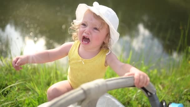 Little funny cute blonde girl child toddler in yellow bodysuit and white hat crying trying climb baby carriage near lake and green grass outside at summer. Childish tantrum. Healthy childhood concept. — Stock Video