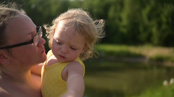 Petit bébé mignon tout-petit fille blonde boucles en combinaison jaune et chapeau blanc sur les bras des mères. Mère embrassant l'enfant à l'extérieur à la campagne l'été. Une famille heureuse saine concept d'enfance. — Video