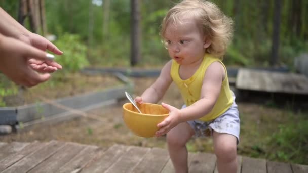 Little funny cute blonde girl child toddler with dirty clothes and face gives dishes to mother after eating baby food fruit or vegetable puree with spoon from yellow plate outside at summer — Stock Video