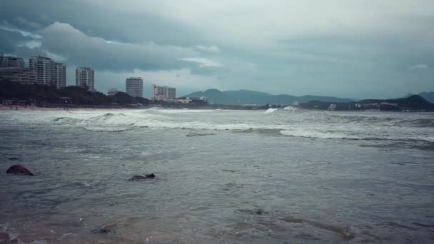 Costa de uma grande e moderna cidade costeira de Sanya, Hainan, com arranha-céus. Ondas surfar em tempo ventoso tempestuoso em cores de água depressivas escuras. Mar sombrio ou oceano . — Vídeo de Stock