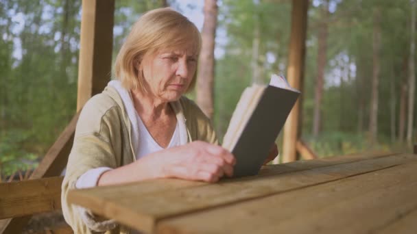 Oudere oudere vrouw met slecht gezichtsvermogen probeert een boek zonder bril te lezen op een houten terras buiten op het zomerse platteland. Vrouwelijke squint van middelbare leeftijd en richt zich op klein tekstlettertype. — Stockvideo