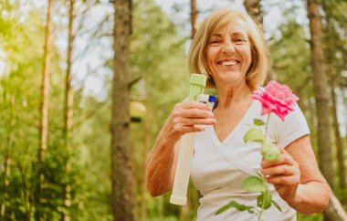 Yaşlı, kıdemli bahçıvan kadın, kır bahçesinde çiçeklere bakan kırsal alanda, su tozu kullanarak çiçek açan bitkileri ilaçlıyor. Çiftçilik, bahçıvanlık, tarım, emekli yaşlılar..