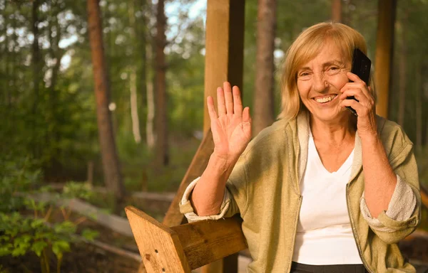 Teknologi, konsep orang tua - wanita tua yang tersenyum bahagia berbicara menggunakan ponsel di luar rumah di teras kayu di taman musim panas di halaman belakang desa — Stok Foto