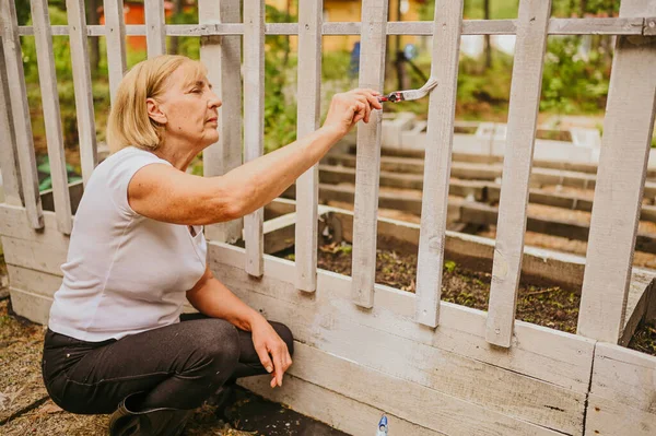 Wanita tukang kebun senior melukis warna putih pagar kayu di pedesaan pertanian musim panas di luar ruangan menggunakan kuas cat. Repaid dan renovasi, pensiunan orang tua yang aktif konsep. — Stok Foto
