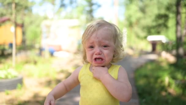 Крупним планом портрет маленької смішної дівчинки-блондинки малюка в жовтому боді костюмі, що плаче надворі влітку. Дитяча істерія. Концепція здорового дитинства . — стокове відео
